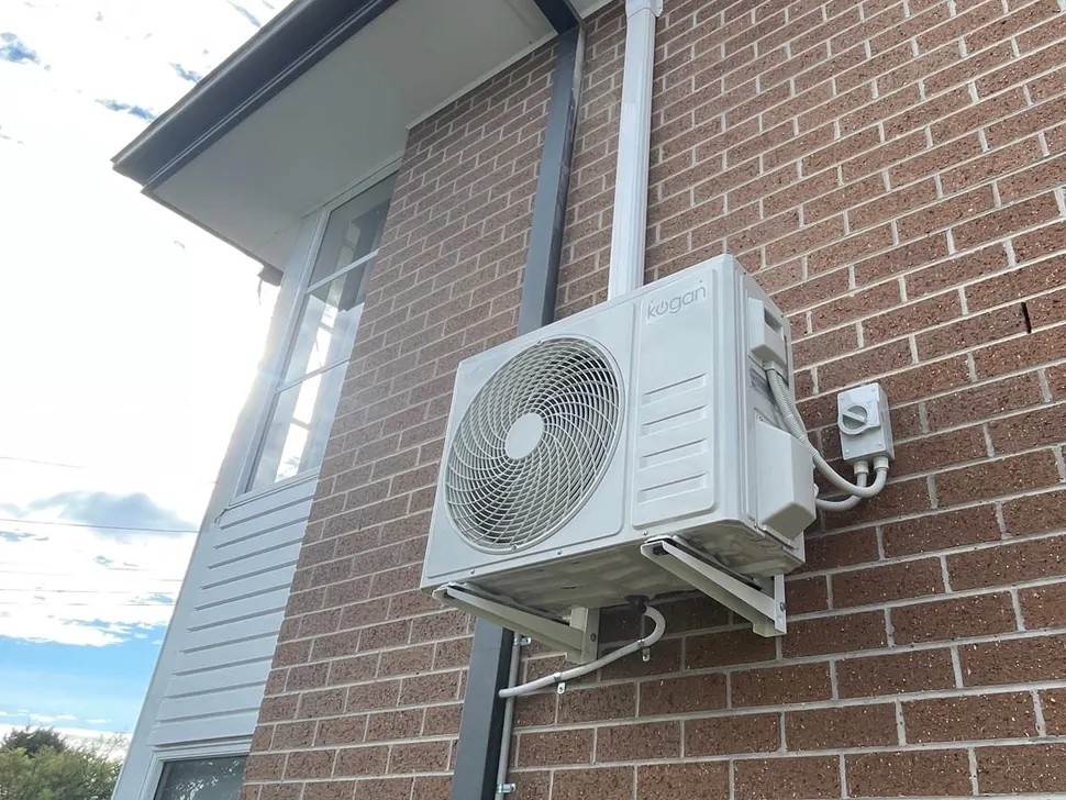 Split system outdoor unit mounted on a wall bracket against a brick wall, with a wall isolating switch, neat conduit and a drain pipe
