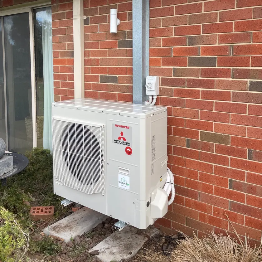 New Mitsubishi Heavy Industries split system outdoor unit standing on pavers against a brick wall, with a pipe cover and a weatherproof isolator above it
