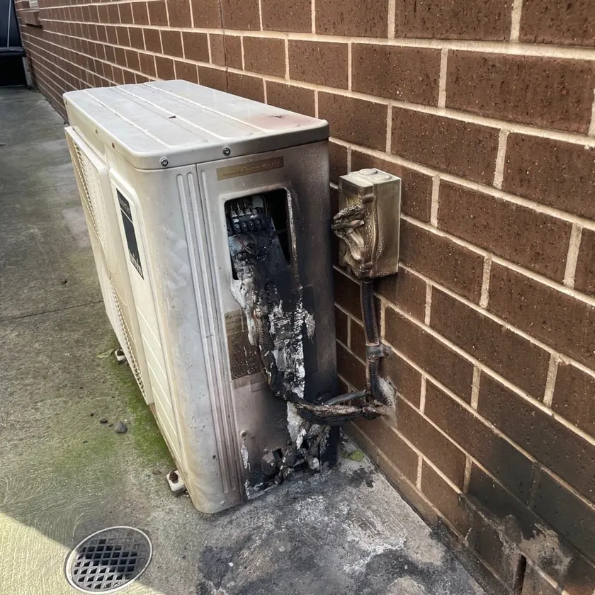 Old split system outdoor unit and isolator burnt out, with scorch marks on the unit, the brick wall and the concrete