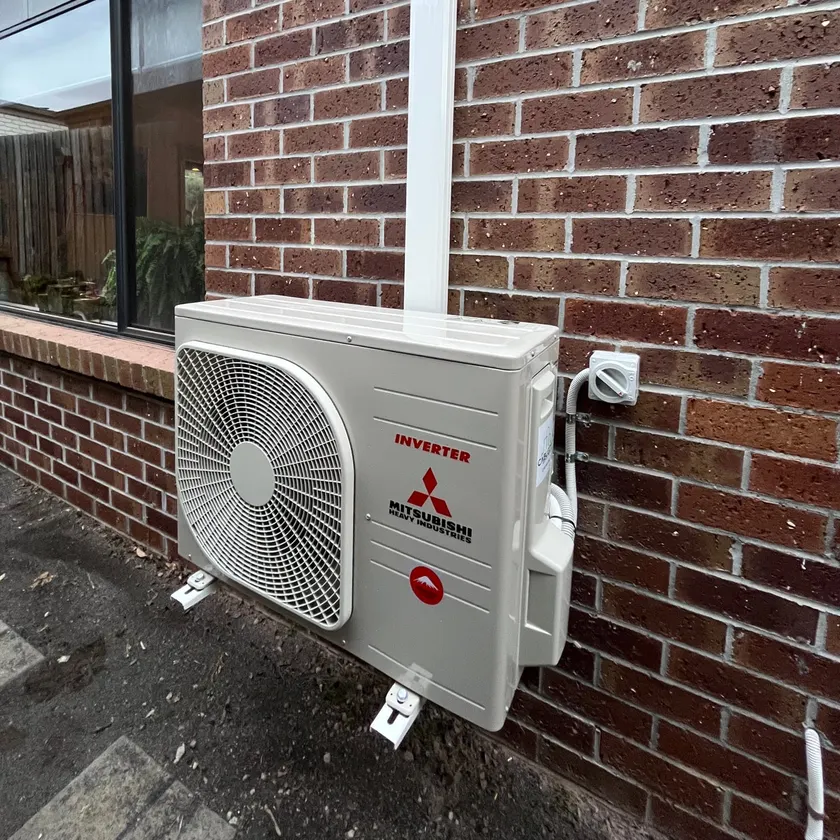 Mitsubishi Heavy Industries split system outdoor unit on mounting feet against a brick wall, with a white pipe cover and an isolator