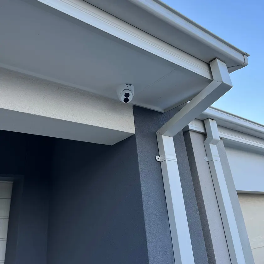 White turret security camera mounted under a white eave against a blue sky