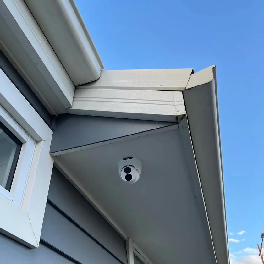 White turret security camera under a corner eave beside weatherboard cladding and windows