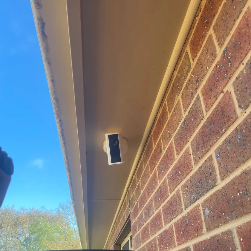 Small white wireless security camera mounted under an eave beside a brick wall