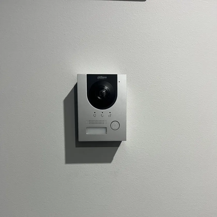 Silver video intercom door station with camera and call button on a white wall
