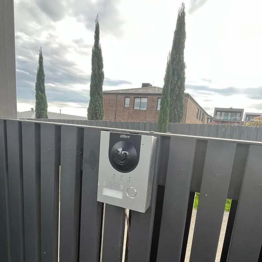 Video door station with camera and call button fixed to a dark slatted fence with cable below