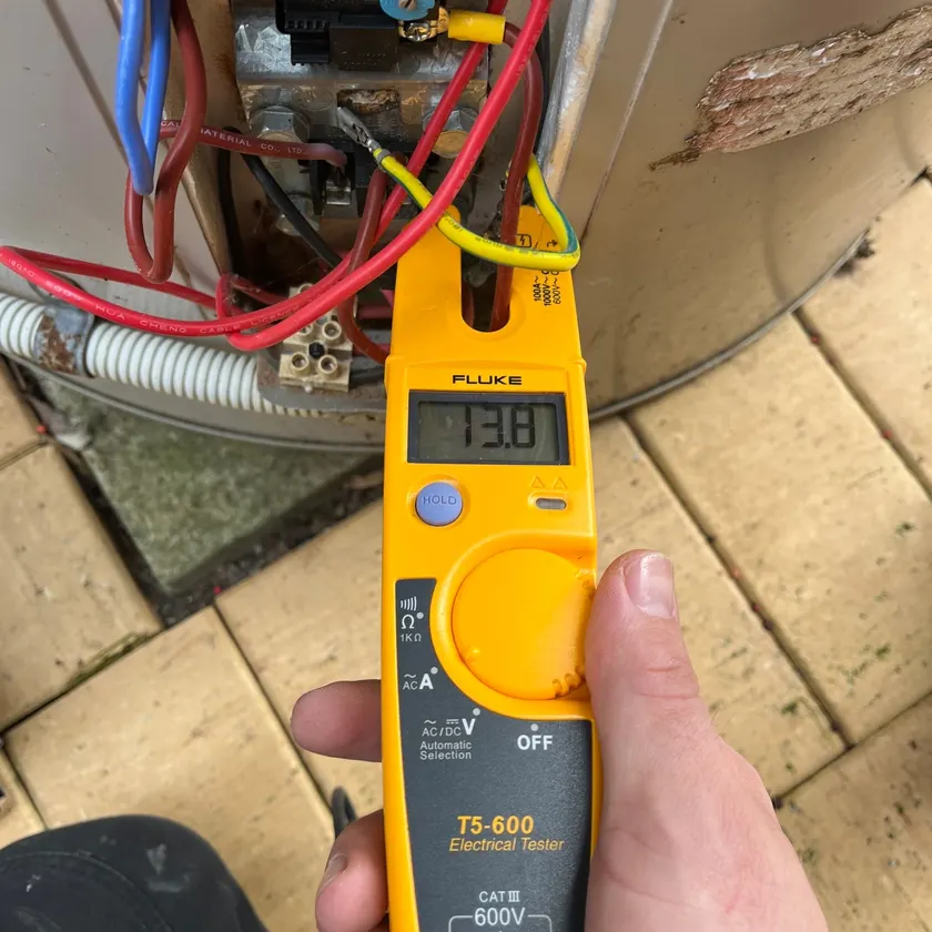 Hand holding an electrical tester showing a reading at an open hot water thermostat cover