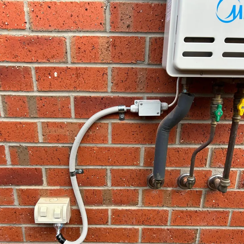 Outdoor hot water unit on a brick wall with a neat flexible conduit run, junction box and weatherproof switch