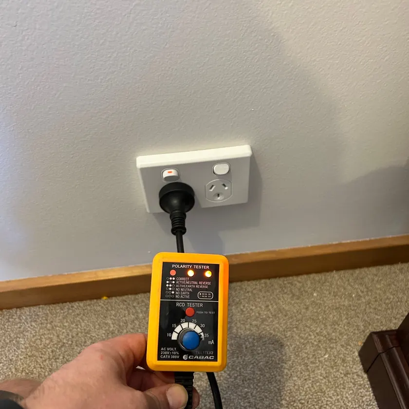 Hand holding a polarity and safety switch tester plugged into a white double power point above a timber skirting