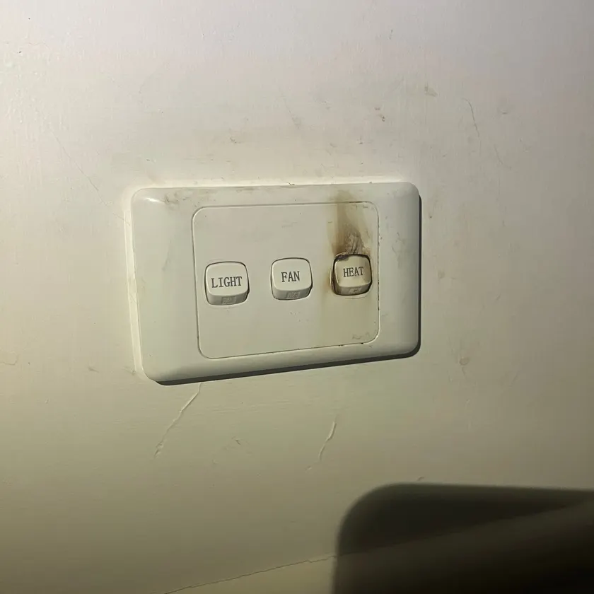 Bathroom light, fan and heat switch plate with brown scorch marks around the heat switch