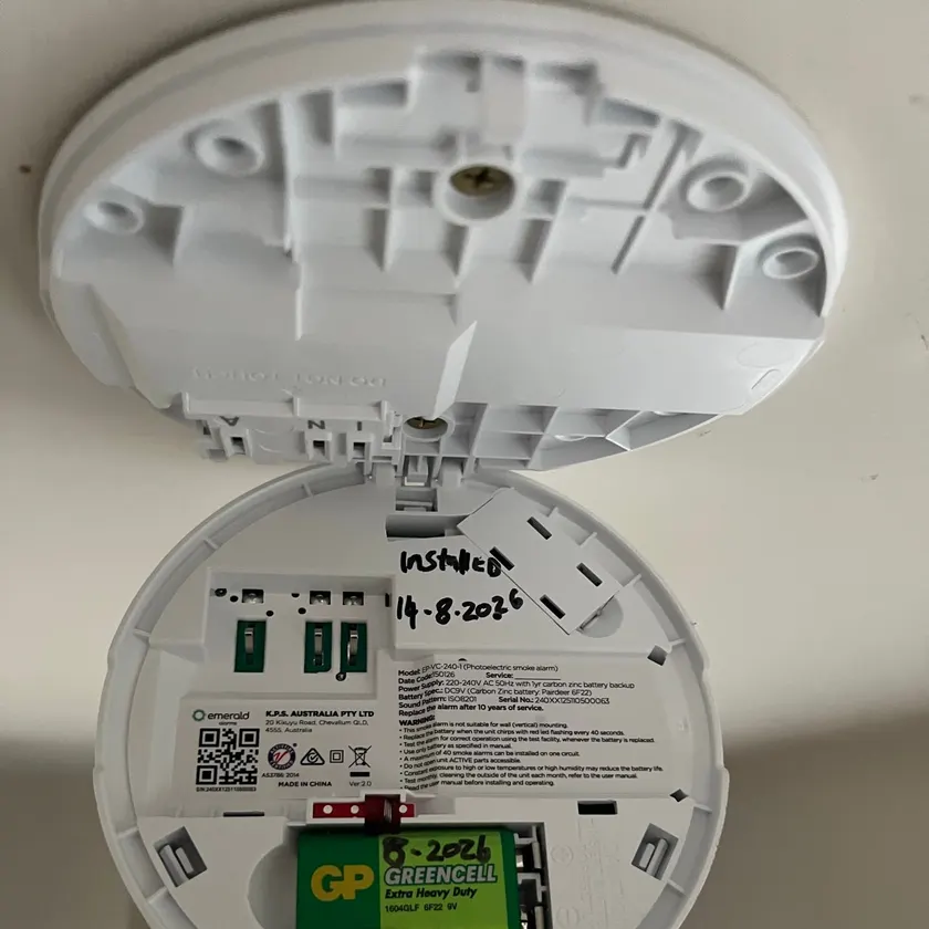 Opened smoke alarm showing a 9V battery and a handwritten Installed 14-8-2026 note