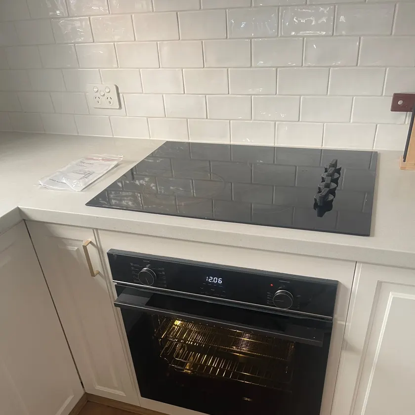 Black wall oven with its light on under a ceramic cooktop in a white kitchen