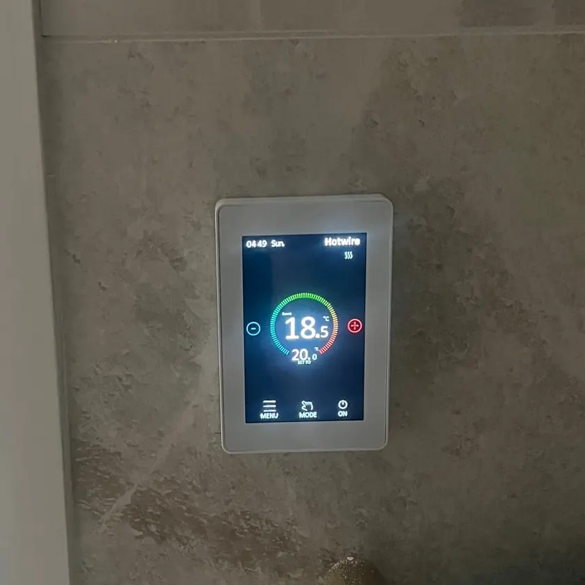 Wall mounted touchscreen thermostat showing 18.5 degrees set to 20 beside a double switch on grey stone-look tiles
