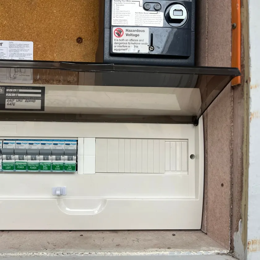 New switchboard with the clear lid open, circuit breakers labelled in order, and a utility hazard sticker