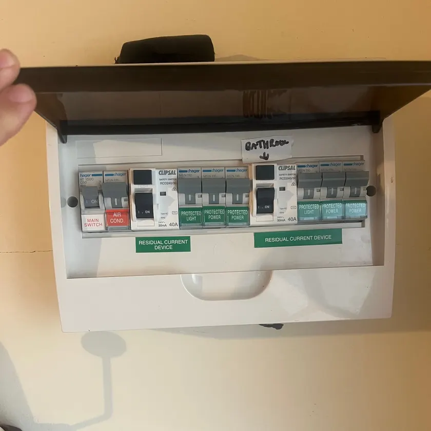 Open white switchboard with two safety switches and labelled circuit breakers held open by a tradie's hand