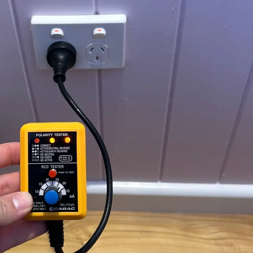 Hand holding a yellow polarity and RCD tester plugged into a white double power point on a panelled wall