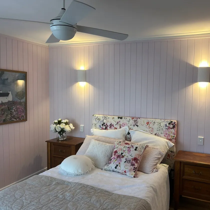 Bedroom with two lit wall lights above a bed, a ceiling fan, and timber panelled walls painted soft pink