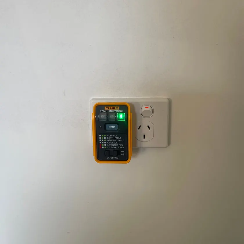 Yellow socket tester with green lights plugged into a white double power point on a plain wall