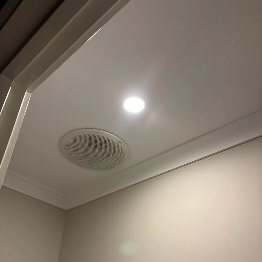 Round exhaust fan grille and a recessed LED downlight on a white ceiling above a cornice
