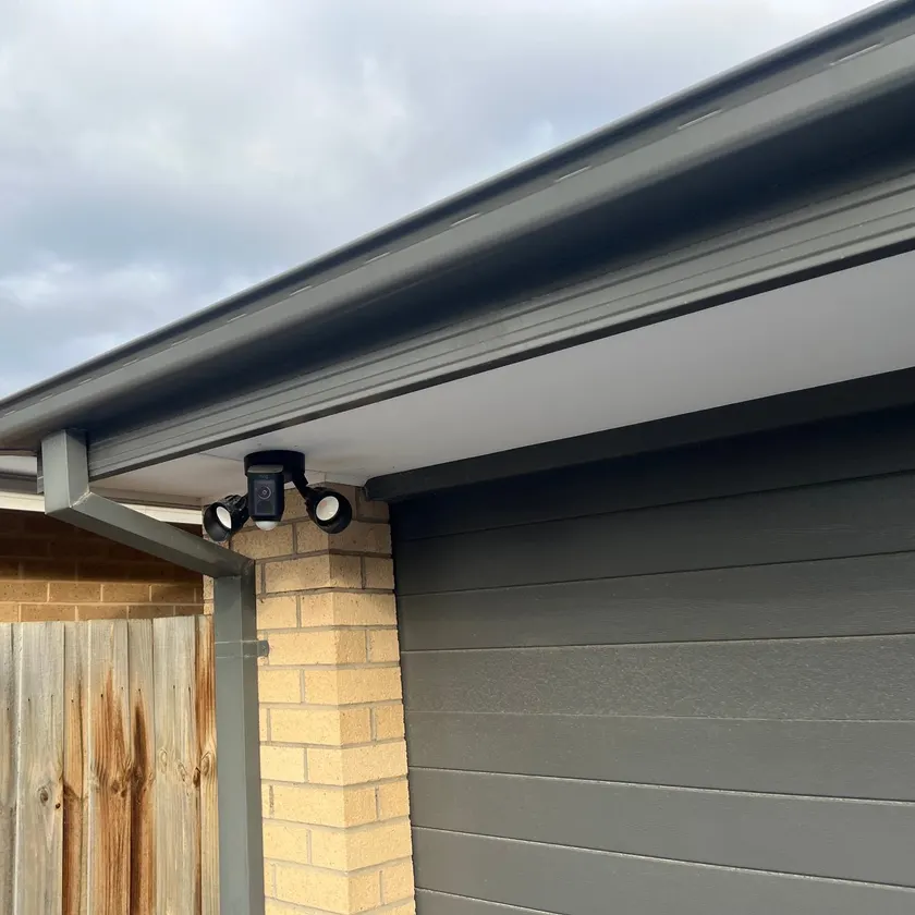 Black twin-head security floodlight with sensor fixed under a garage eave beside a downpipe