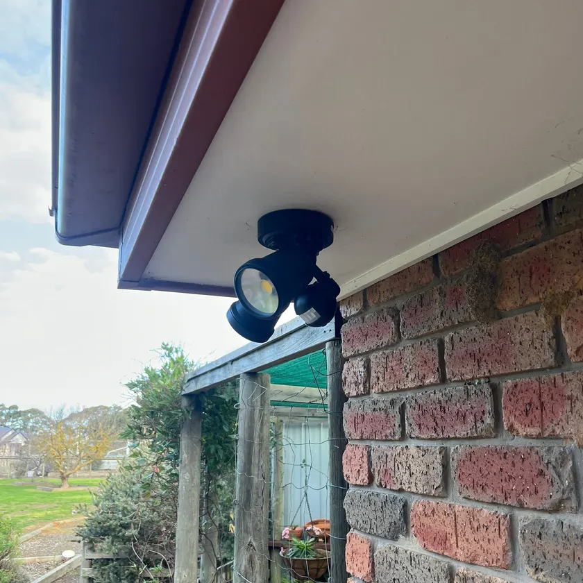 Black twin-head LED sensor floodlight fixed under the eave of a brick house with garden behind