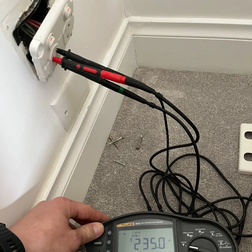 Hand holding a multifunction tester reading 235 volts with probes in a power point