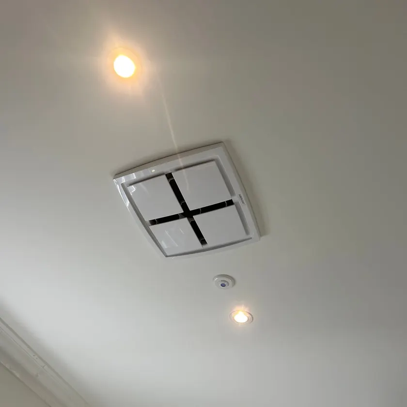 Square white exhaust fan between two lit downlights in a ceiling