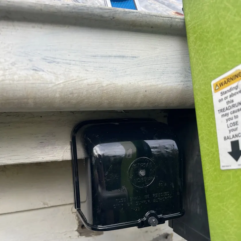 New black service fuse box fitted under the eave of a weatherboard house, ladder beside it