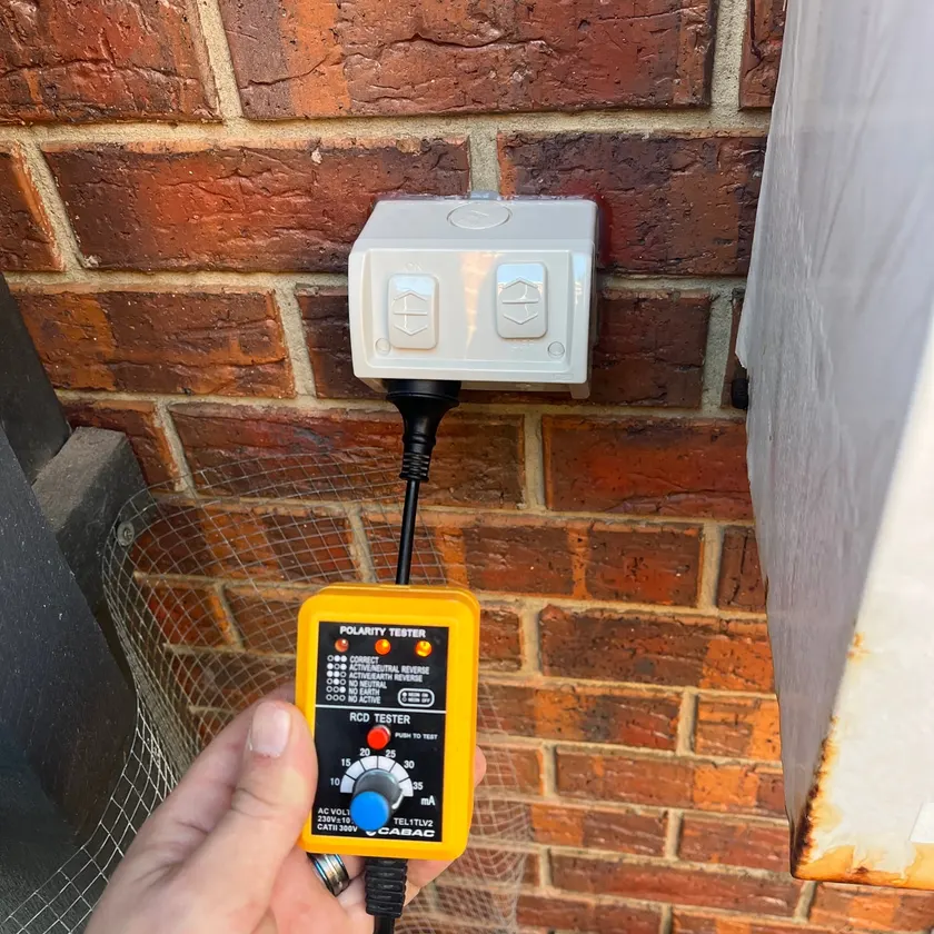 Hand holding a tester plugged into a new weatherproof double power point on a red brick wall