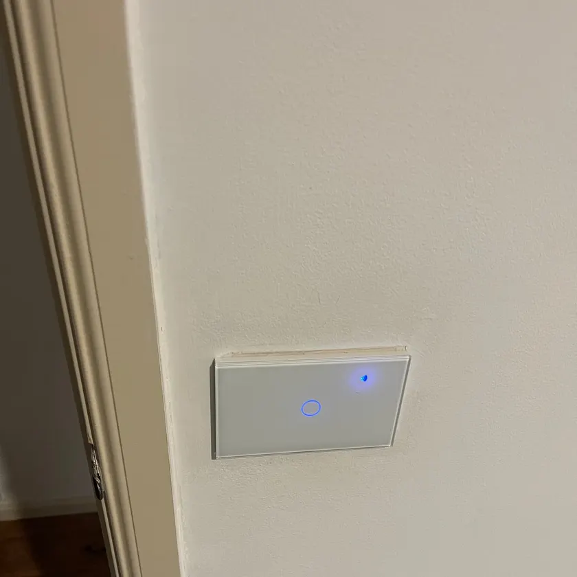 Single-gang white glass smart touch switch with a blue indicator light on a painted wall beside a door frame