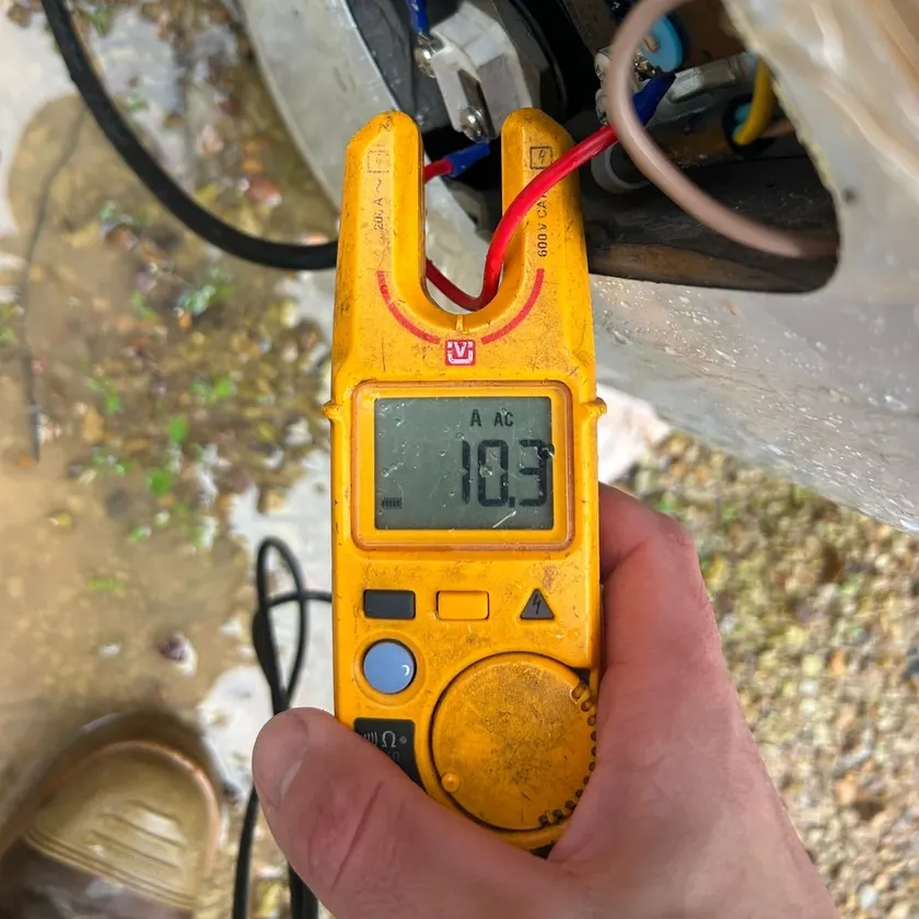 Hand holding a clamp meter reading 10.3 amps on a hot water unit's wiring